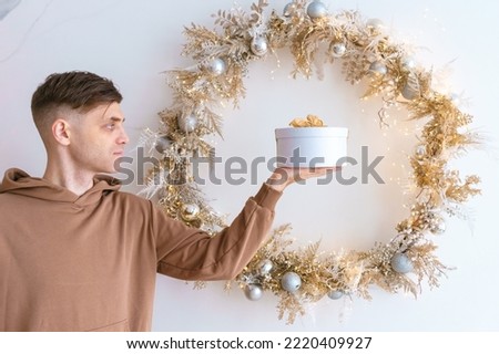 Happy young man holding gift box on background Christmas wreath on background of a light wall at home, shows a hand gesture. Festive mood and promotion of gifts