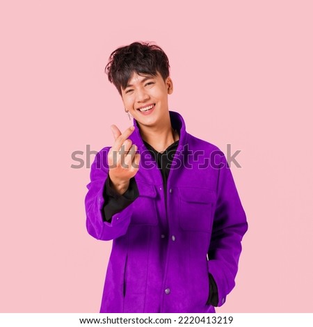 Portrait of a happy Asian handsome man in fashionable purple jacket clothing standing smiling using fingers to make a mini heart sign isolated on pink background. young male fashion model lifestyle.