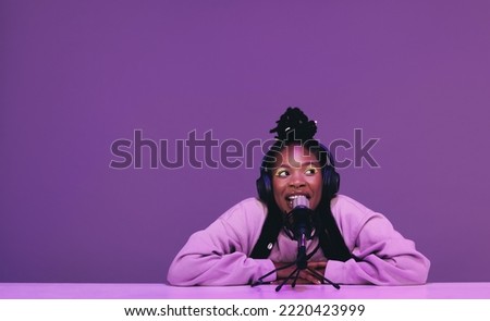 Female podcaster speaking into a microphone while sitting against a purple background. Happy woman recording a live audio broadcast in a studio. Young woman creating content for her internet podcast.