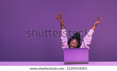 Happy female gamer celebrating winning a video game in a studio. Cheerful young woman playing an online game with a laptop and headsets. Young woman streaming a game against a purple background.
