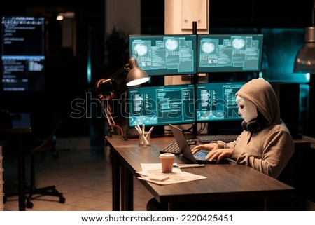 Scammer with mask and hood hacking security server, installing virus to create computer malware and steal big online data. Masked criminal hacker breaking netowrk system at night. Royalty-Free Stock Photo #2220425451