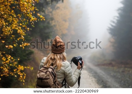 Woman with camera and backpack hiking in misty autumn forest. female tourist photographing nature Royalty-Free Stock Photo #2220430969
