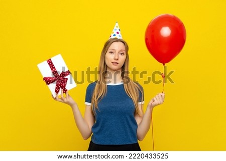 Sad young woman holds box with gift present and red inflated helium balloon, has bad mood during celebration, wears party cone and blue t shirt, feels embarrassed
