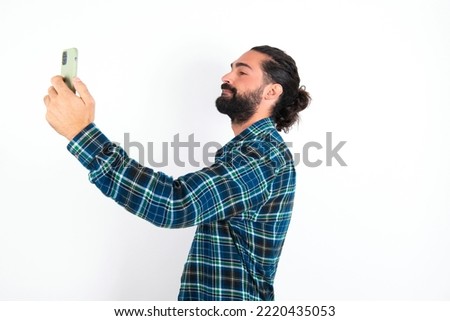 Portrait of a young hispanic bearded man wearing plaid shirt over white background taking a selfie to send it to friends and followers or post it on his social media.