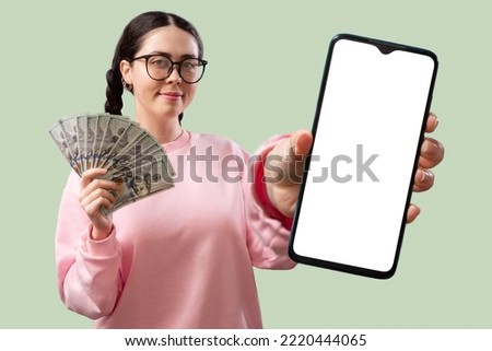 Attractive young Caucasian woman with glasses shows a smartphone with a white screen and holds a fan of money on a light green background. Mock up. The concept of bank app and investments.