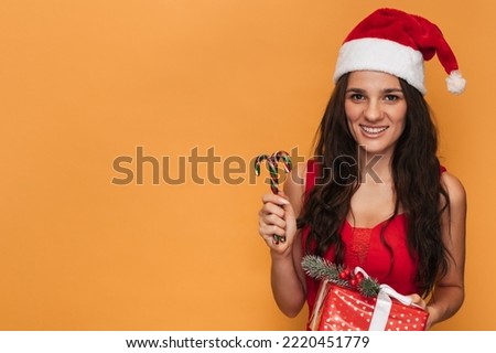 A smiling Caucasian woman in a red Santa hat with a gift box in her hands on a yellow isolated background. The concept of Christmas and New Year. A place for your text.