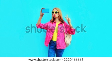 Portrait of happy stylish young woman taking selfie with smartphone blowing her lips sends kiss wearing pink jacket, backpack, yellow sunglasses on blue background