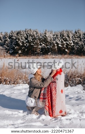Mom and daughter in white knitted hats play in the snow near the lake and forest. Mom wrapped her daughter in a red blanket for a walk cold winter.