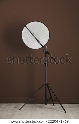 Studio reflector on tripod near brown wall indoors. Professional photographer's equipment