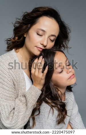 brunette woman with closed eyes embracing head of daughter isolated on grey