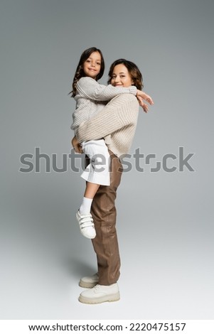 happy and trendy woman holding smiling daughter in hands while looking at camera on grey background
