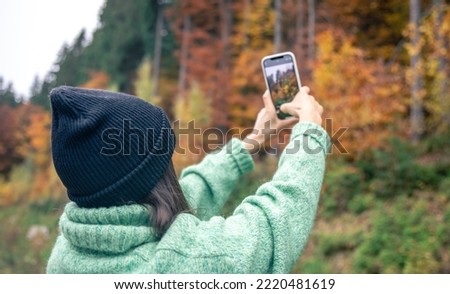 A young woman in a black hat takes a photo of the autumn forest on a smartphone.