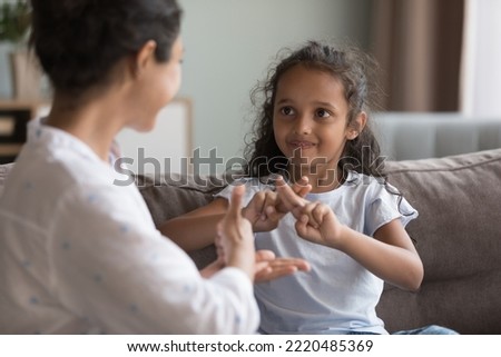Indian preschooler girl and young mother showing symbols with hands using visual-manual gestures enjoy communication seated on couch at home. Sign language usage