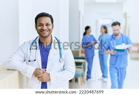 Smiling Indian doctor standing in hallway of modern hospital against background of working medical assistants and nurses. Medical institution