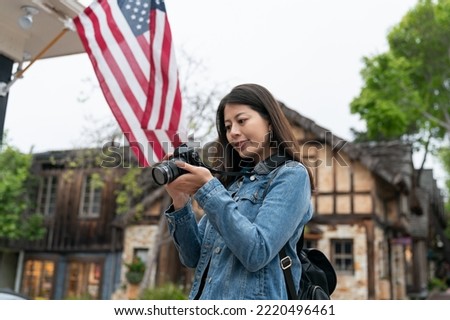 curious asian woman visitor looking into distance with camera in hand while photographing street scenes of the fairytale town Carmel by the sea against America flag background