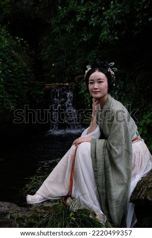 A beautiful Asian lady in green vintage dress sitting on a rock with head resting on one hand looking at camera before waterfall background 