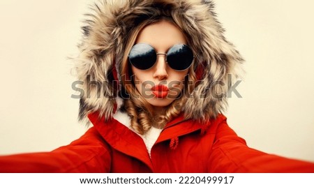 Portrait of stylish woman blowing her lips sends air kiss stretching hand for taking selfie with smartphone wearing jacket with fur hood