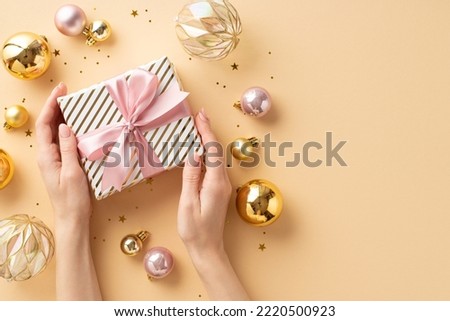 New Year concept. First person top view photo of female hands holding present box with pink ribbon bow over transparent gold pink baubles confetti on isolated pastel beige background with empty space