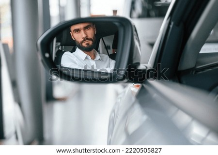 Looking in the side mirror. Young man in white clothes is in the car dealership.