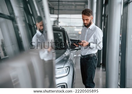 Black graphic tablet in hands. Young man in white clothes is in the car dealership.