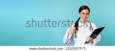 Confident smiling doctor woman, physician showing thumbs up, holding clipboard, appointment in hospital, standing over torquoise background.