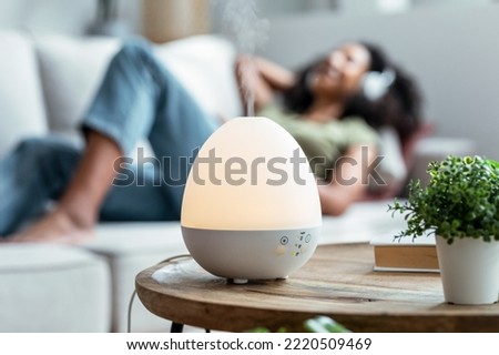 Shot of essential oil aroma diffuser humidifier diffusing water articles in the air while woman listening music lying on coach. Royalty-Free Stock Photo #2220509469