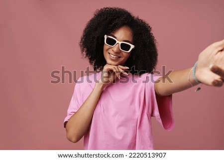 Selfie of young beautiful smiling curly woman in sun glasses touching her chin, while standing over isolated background