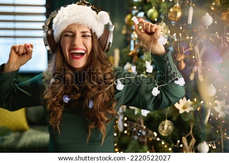 Christmas time. happy stylish middle aged woman with Santa hat in green dress listening to the music with headphones and dancing near Christmas tree in the modern house.