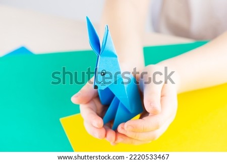 Children's craft origami blue rabbit. Origami blue paper rabbiton a table in the hands of a child. Symbol of the year.