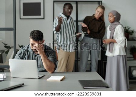Middle aged Hispanic businessman sitting by workplace and networking against group of cruel intercultural colleagues bullying him Royalty-Free Stock Photo #2220559831