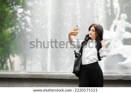 Beautiful brunette in a white shirt and black pants makes a selfie on the phone on the background of a fountain