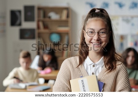 Smiling youthful schoolgirl in eyeglasses and casualawear looking at camera while standing in front of her classmates at lesson Royalty-Free Stock Photo #2220575553