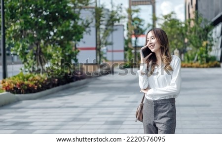 Smiling Asian curly business woman wearing trendy walks down the central city street and uses her phone. Pretty summer woman in white shirt walks down the street looking at her mobile phone