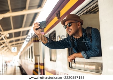 Young asian man travel by train. Sticking his head outta the train window taking photo. Explorer Backpacker arrival and departure at platform railway. Freedom  trip on vacation time holiday weekend.