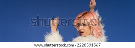 woman in white and fluffy faux fur jacket looking at camera while dancing isolated on blue, banner