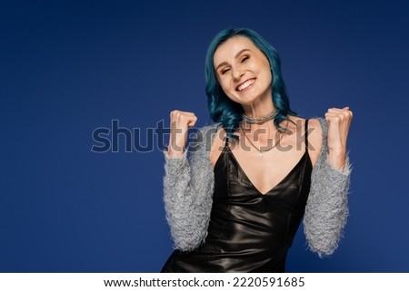excited woman in black leather jacket and faux fur sleeves showing win gesture isolated on blue