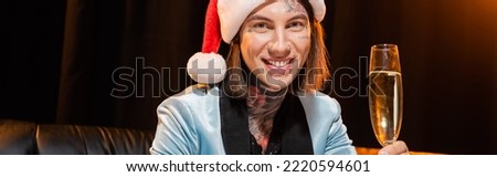 smiling tattooed queer person in santa hat looking at camera near champagne glass on dark background, banner