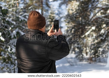 Back view of a man in a black jacket and brown hat who walks in a winter snowy forest on a sunny day and takes pictures and videos on a mobile phone camera, copy space.