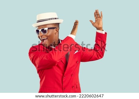 Young energetic African American man wearing sunglasses and red entertainment suit, yelling and clapping hands performing funny dance or working as host of party stands on pastel blue background Royalty-Free Stock Photo #2220602647