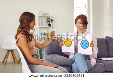 Friendly female child psychologist talks to teenage girl about emotions she is currently feeling. Psychologist shows teenage girl two sheets of paper with positive and sad emoticons drawn on them.