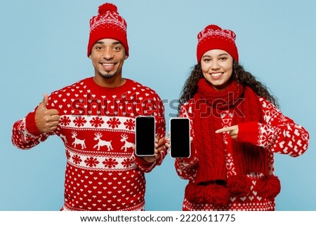 Merry young couple two man woman wear red Christmas sweater Santa hat posing hold use mobile cell phone with blank screen show thumb up isolated on plain blue background. Happy New Year 2023 concept