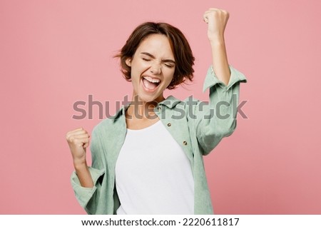 Young happy woman 20s she wear green shirt white t-shirt doing winner gesture celebrate clenching fists say yes isolated on plain pastel light pink background studio portrait. People lifestyle concept Royalty-Free Stock Photo #2220611817