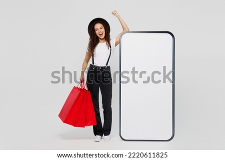 Full body young woman wear t-shirt hat hold shopping red paper package bag big blank screen mobile phone with area do winner gesture isolated on plain white background. Black Friday sale buy concept