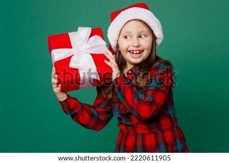 Merry wistful fun cute little child kid girl 6-7 years old wear red dress Christmas hat posing hold present box with gift ribbon bow isolated on plain dark green background Happy New Year 2023 concept