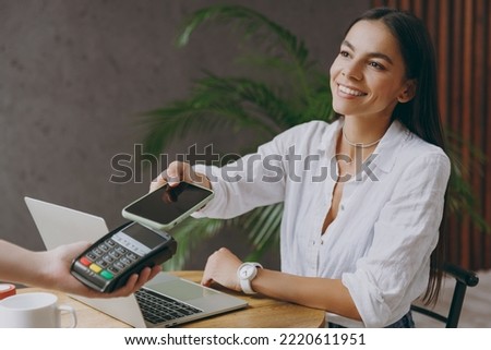 Young happy woman wear white shirt hold pay waiter hold mobile cell phone wireless bank payment terminal waiter sit at table in coffee shop cafe restaurant indoor work or study on laptop pc computer Royalty-Free Stock Photo #2220611951