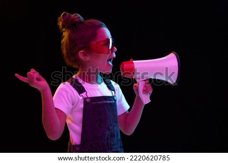 Closeup. Stylish little girl, pupil in white t-shirt and denim dress shouring at megaphone isolated over dark background in neon light. Concept of beauty, kids fashion, children emotions. Royalty-Free Stock Photo #2220620785