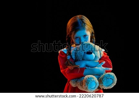 Sadness. Little girl with pigtails wearing red festive dress standing with teddy bear isolated over dark background. Concept of children emotions, child psychology