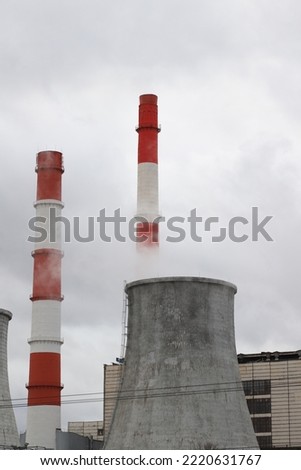 Cooling towers and red-white pipes of the city's thermal power plant against the background of a gray sky and white puffs of steam.