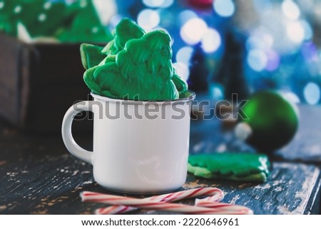 Frosted Christmas tree shape cookies or biscuits with green icing inside of a white coffee cup with candy canes nearby. Selective focus with blurred foreground and background.