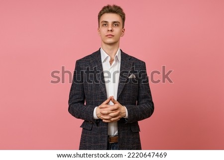 young businessman in a jacket and white shirt with his palms crossed in front of him proudly looking at the camera on a pink background